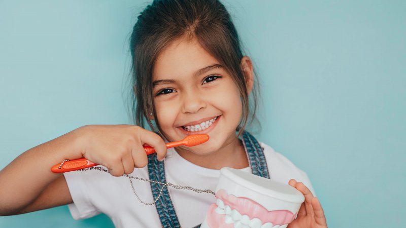 Young girl smiling and brushing her teeth while holding a teeth model