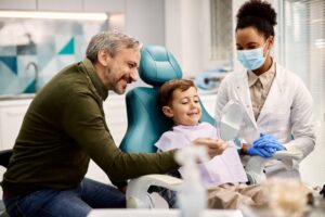 Father and child at dentist, child in dentist's chair with dentist looking on. 